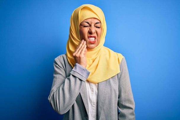 Keski-ikä brunette liiketoiminnan nainen yllään muslimi perinteinen hijab yli sininen tausta koskettaa suuhun käsin kivulias ilme, koska hammassärky tai hampaiden sairaus hampaita. Hammaslääkäri
 - Valokuva, kuva