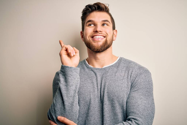 Young handsome blond man with beard and blue eyes wearing casual sweater with a big smile on face, pointing with hand and finger to the side looking at the camera. - Photo, image