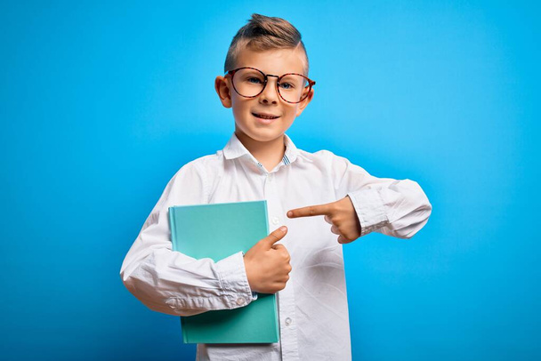 Joven niña estudiante caucásica usando gafas inteligentes y sosteniendo un libro de la escuela muy feliz señalando con la mano y el dedo
 - Foto, imagen