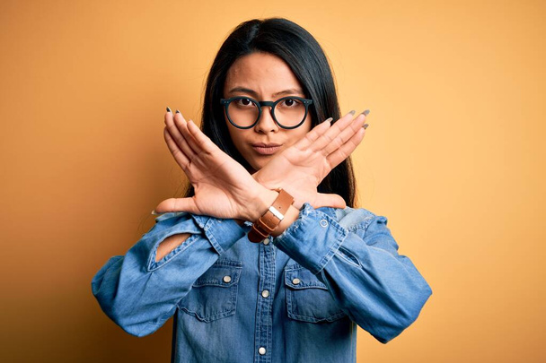 Young beautiful chinese woman wearing casual denim shirt over isolated yellow background Rejection expression crossing arms doing negative sign, angry face - Φωτογραφία, εικόνα