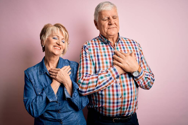 Senior kaunis pari seisoo yhdessä yli eristetty vaaleanpunainen tausta hymyilee kädet rinnassa suljetuin silmin ja kiitollinen ele kasvot. Terveyskäsite
. - Valokuva, kuva