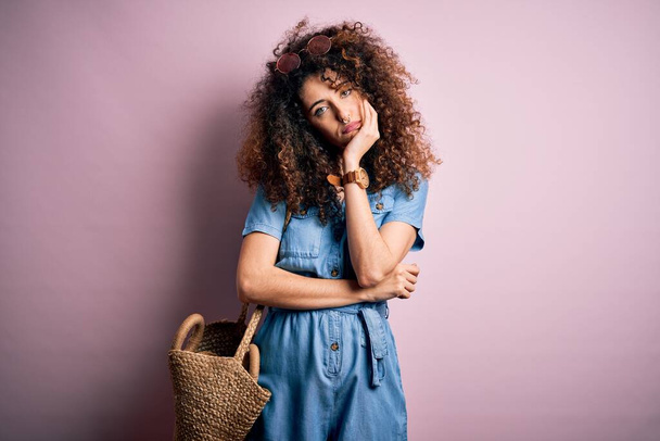 Young beautiful woman with curly hair and piercing wearing denim dress and wicker bag thinking looking tired and bored with depression problems with crossed arms. - Foto, Imagen