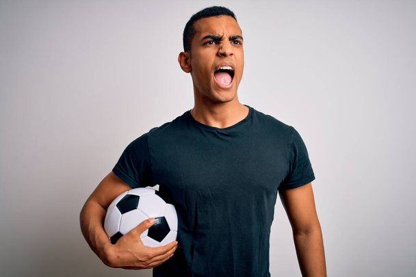 Hombre afroamericano guapo jugando fútbol sosteniendo pelota sobre fondo blanco enojado y loco gritando frustrado y furioso, gritando con ira. Rabia y concepto agresivo
. - Foto, Imagen