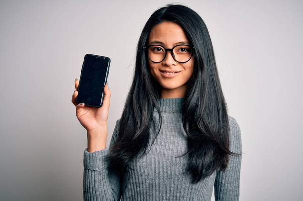 Young beautiful chinese woman holding smartphone over isolated white background with a happy face standing and smiling with a confident smile showing teeth - Photo, Image