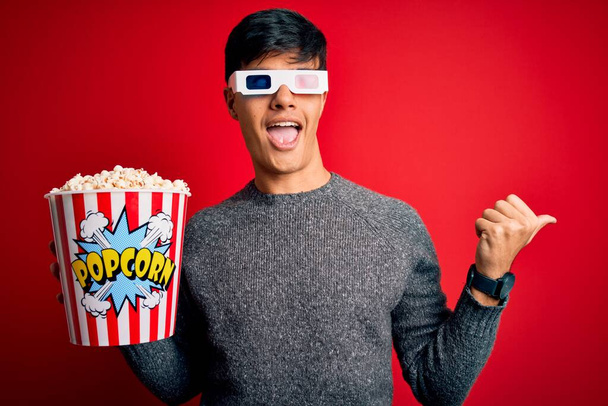 Young handsome man watching movie eating popcorns snack over isolated red background pointing and showing with thumb up to the side with happy face smiling - Photo, Image