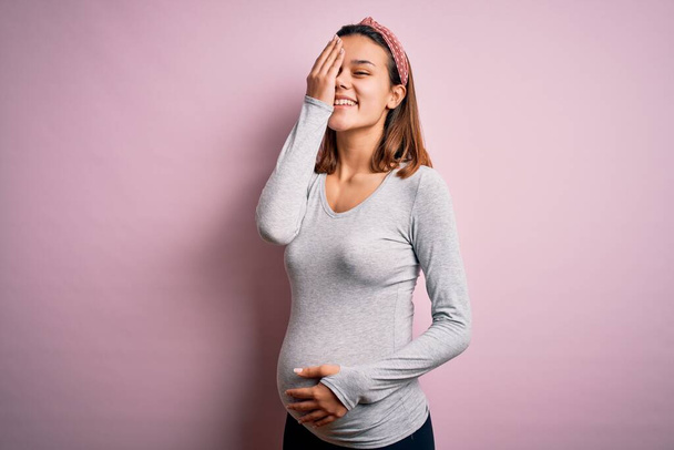Young beautiful teenager girl pregnant expecting baby over isolated pink background covering one eye with hand, confident smile on face and surprise emotion. - 写真・画像