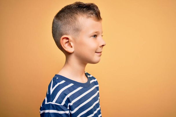 Junge kleine kaukasische Kind mit blauen Augen trägt nautische gestreiftes Hemd über gelbem Hintergrund zur Seite schauen, entspannen Profil Pose mit natürlichem Gesicht mit selbstbewusstem Lächeln. - Foto, Bild