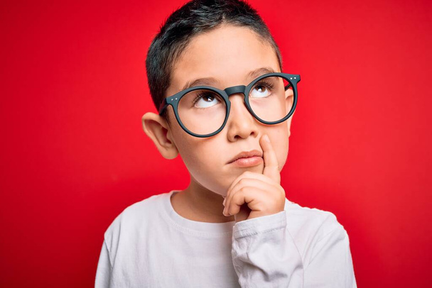İnek gözlüğü takan küçük zeki çocuk kırmızı izole edilmiş arka planda ciddi bir yüz soru düşünüyor, çok karışık bir fikir. - Fotoğraf, Görsel