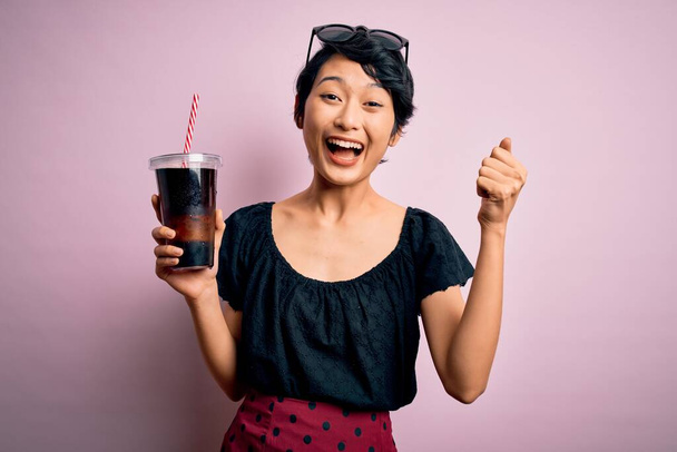 Genç, güzel Çinli bir kadın pembe arka planda saman kullanarak kola içiyor. Gururla bağırıyor, zaferi ve başarıyı kutluyor. Çok heyecanlı, coşkulu bir duygu. - Fotoğraf, Görsel