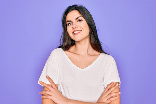 Jeune belle femme brune portant un t-shirt blanc décontracté sur fond violet heureux visage souriant avec les bras croisés regardant la caméra. Personne positive
. - Photo, image