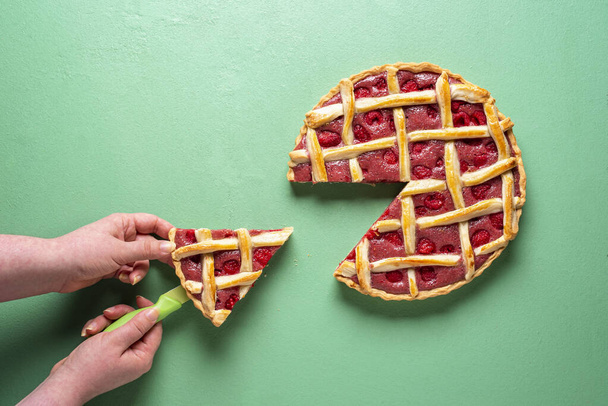 Woman's grabbing slice of raspberry tart, above view on a green background. Sweet pie home-baked with organic raspberries. Traditional lattice crust fruits filling pie above view. - Photo, Image