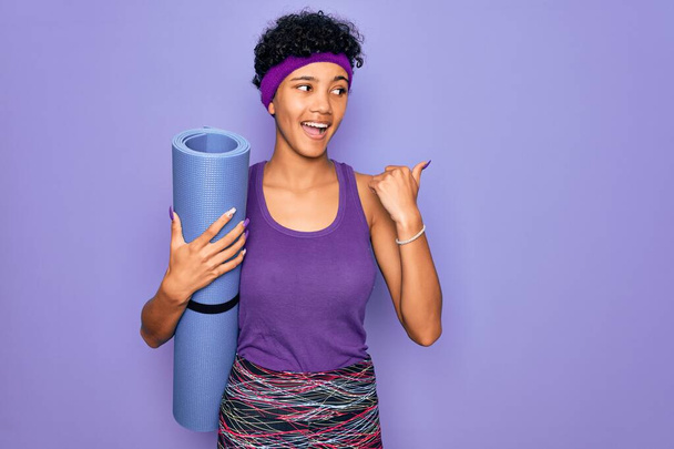Молодая красивая африканская спортивная женщина в спортивной одежде держит коврик для йоги указывая и показывая с большим пальцем в сторону со счастливым лицом улыбаясь
 - Фото, изображение