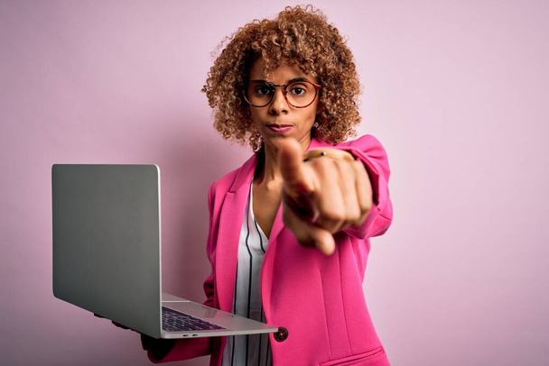Afroamerykanka biznes kobieta z kręconymi włosami pracuje przy użyciu laptopa na różowym tle wskazując palcem na aparat i do Ciebie, znak ręczny, pozytywny i pewny gest z przodu - Zdjęcie, obraz