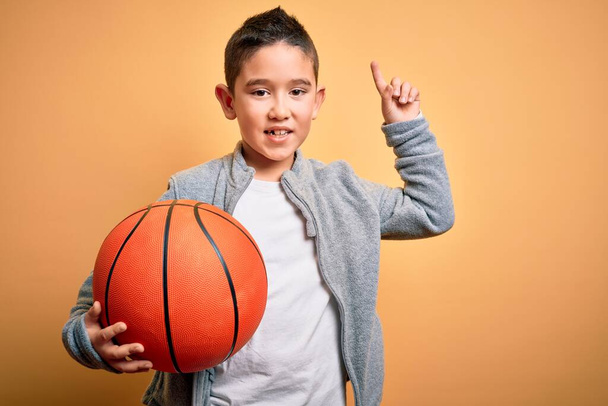 Mladý chlapec kluk hraje s basketbalovým míčem na izolovaném žlutém pozadí překvapen nápadem nebo otázkou ukazující prstem se šťastnou tváří, číslo jedna - Fotografie, Obrázek