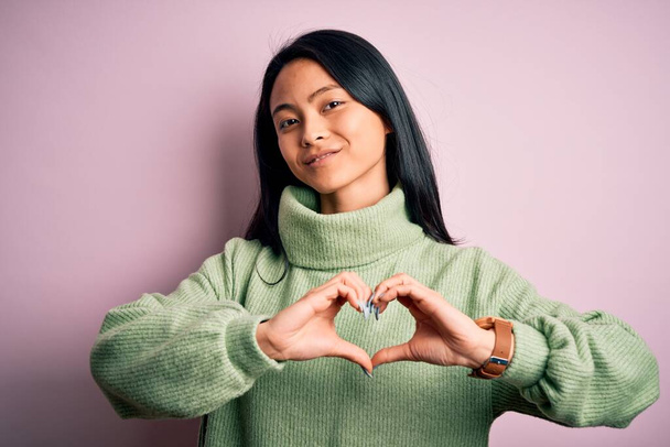 Young beautiful chinese woman wearing turtleneck sweater over isolated pink background smiling in love showing heart symbol and shape with hands. Romantic concept. - Photo, Image