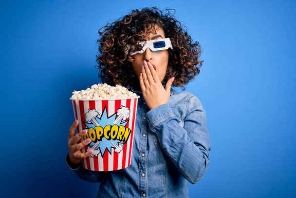Young beautiful curly arab woman watching movie using 3d glasses eating popcorn snack cover mouth with hand shocked with shame for mistake, expression of fear, scared in silence, secret concept - Фото, изображение