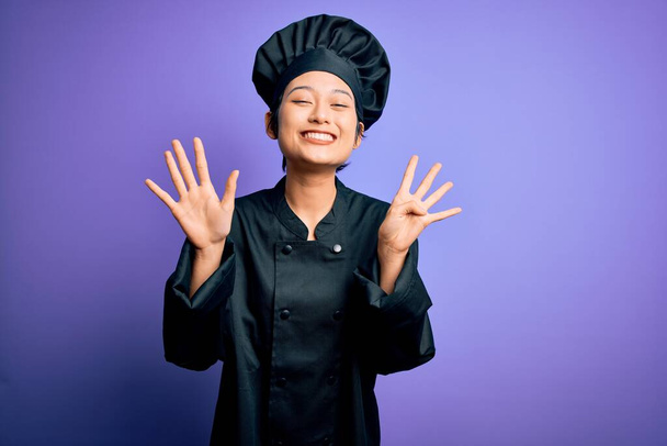 若いです美しいです中国人シェフ女性身に着けています炊飯器の制服と帽子以上紫の背景表示と指番号9で指摘しながら、自信と幸せ. - 写真・画像