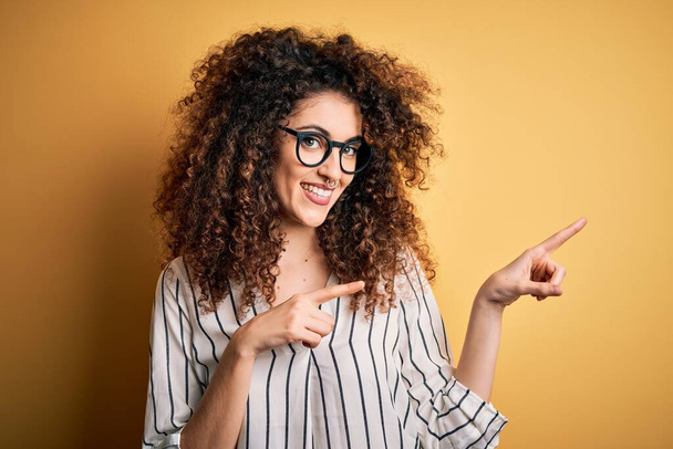 Young beautiful woman with curly hair and piercing wearing striped shirt and glasses smiling and looking at the camera pointing with two hands and fingers to the side. - Photo, Image