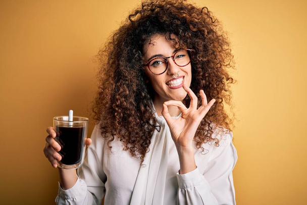 Nuori kaunis nainen kihara tukka ja lävistyksiä yllään lasit juominen lasi kahvia tekee ok merkki sormilla, erinomainen symboli
 - Valokuva, kuva