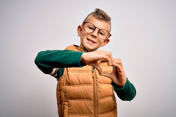 Ein kleines kaukasisches Kind mit blauen Augen, Wintermantel und schicker Brille, das verliebt lächelt und mit den Händen Herzsymbole in Form bringt. Romantisches Konzept. - Foto, Bild