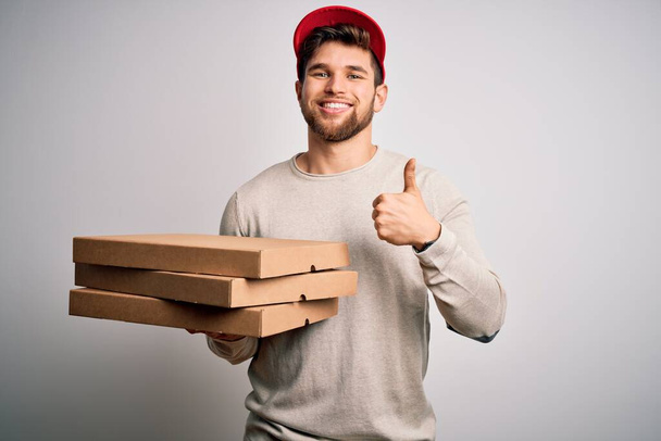 Young blond delivery man with beard and blue eyes holding cardboards of pizza happy with big smile doing ok sign, thumb up with fingers, excellent sign - Photo, Image