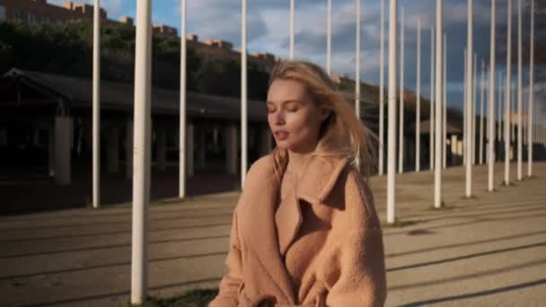 Średnie ujęcie romantycznej blondynki w płaszczu pewnie spacerującej ulicą o zachodzie słońca - Materiał filmowy, wideo