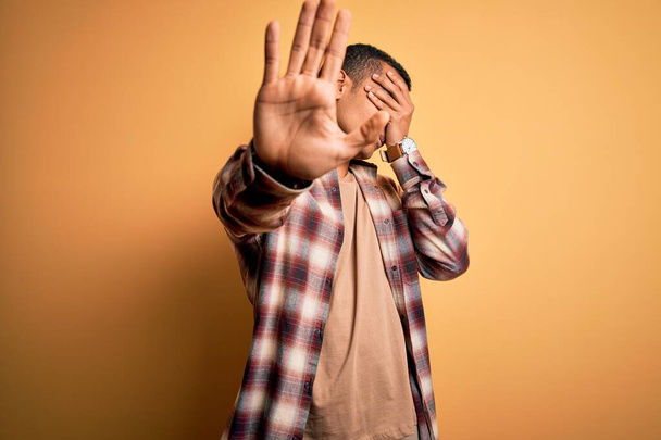 Young handsome african american man wearing casual shirt standing over yellow background covering eyes with hands and doing stop gesture with sad and fear expression. Embarrassed and negative concept. - Valokuva, kuva