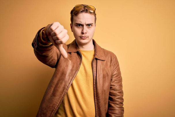 Молодой красивый рыжий мужчина в кожаной куртке на изолированном желтом фоне, выглядящий несчастным и злым, демонстрирующий отказ и негатив с опущенными пальцами. Плохое выражение
. - Фото, изображение