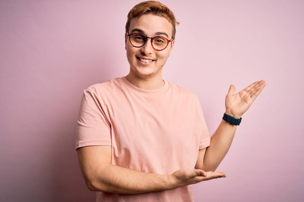 Joven hombre pelirrojo guapo con camiseta casual de pie sobre fondo rosa aislado invitando a entrar sonriente natural con la mano abierta
 - Foto, imagen