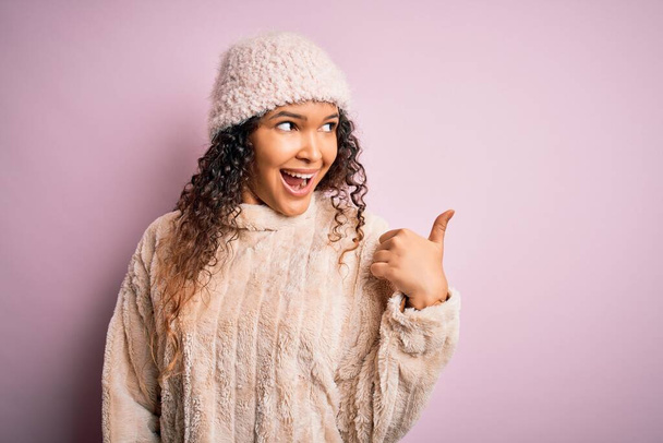 Beautiful woman with curly hair wearing casual sweater and wool cap over pink background smiling with happy face looking and pointing to the side with thumb up. - Photo, image