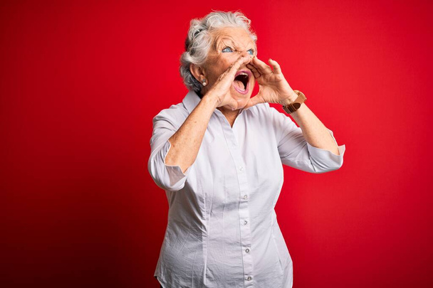 Senior kaunis nainen yllään tyylikäs paita seisoo yli eristetty punainen tausta huutaa vihainen ääneen kädet suun yli
 - Valokuva, kuva