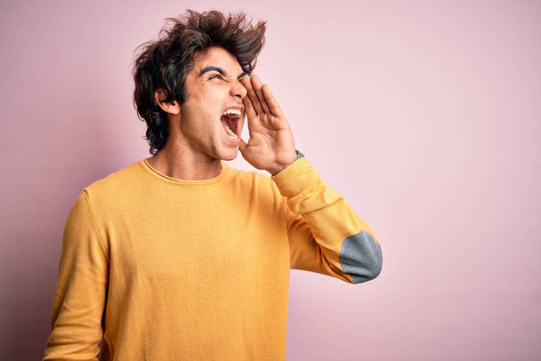 Homem bonito jovem vestindo camiseta casual amarela em pé sobre fundo rosa isolado gritando e gritando alto para o lado com a mão na boca. Conceito de comunicação
. - Foto, Imagem
