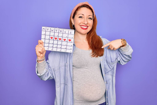 Junge schöne rothaarige schwangere Frau erwartet Baby Wartezeit Kalender mit Überraschung Gesicht zeigt mit dem Finger auf sich selbst - Foto, Bild