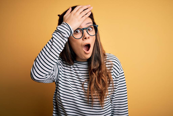 Young beautiful brunette woman wearing french beret and glasses over yellow background surprised with hand on head for mistake, remember error. Forgot, bad memory concept. - Foto, Imagen