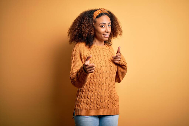 Jovem americana africana com cabelo afro vestindo camisola casual sobre fundo amarelo apontando os dedos para a câmera com rosto feliz e engraçado. Boa energia e vibrações
. - Foto, Imagem