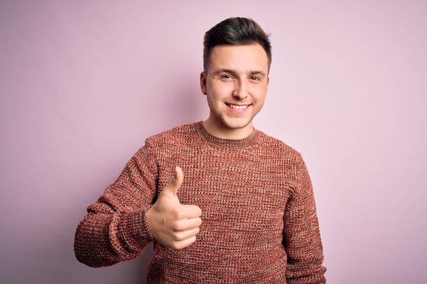 Jonge knappe blanke man draagt casual winter trui over roze geïsoleerde achtergrond doet vrolijke duimen omhoog gebaar met de hand. Goedkeurende uitdrukking die naar de camera kijkt en succes laat zien. - Foto, afbeelding