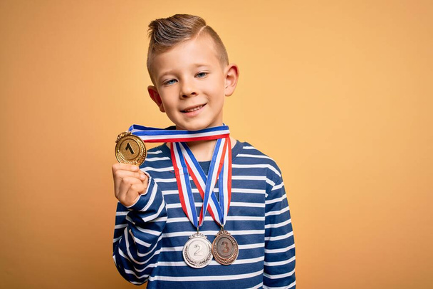 若い白人の勝者の子供は黄色の背景に賞のメダルを身に着けています幸せな顔立ちと笑顔で自信を持って笑顔歯を示す - 写真・画像