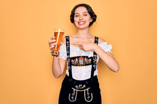 Giovane bella donna tedesca con gli occhi azzurri che indossa l'abito octoberfest bere bicchiere di birra molto felice puntando con mano e dito
 - Foto, immagini