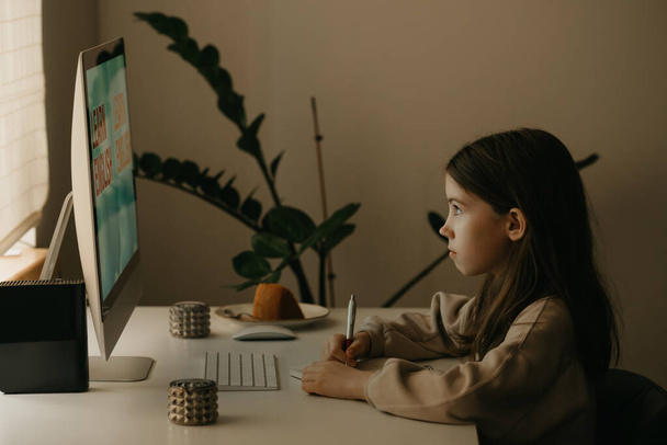 Afstandsonderwijs. Een jong meisje met lang haar dat op afstand online studeert. Een vrouwelijk kind leert thuis een les met een alles-in-één computer. Thuisonderwijs. - Foto, afbeelding
