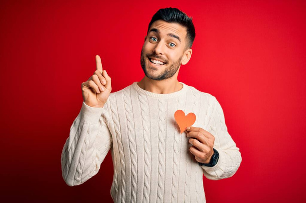 Nuori komea mies, jolla on punainen muoto sydän romanttisena ja terveydellisenä symbolina yllättynyt ideasta tai kysymyksestä, joka osoittaa sormella onnellisia kasvoja, numero yksi.
 - Valokuva, kuva
