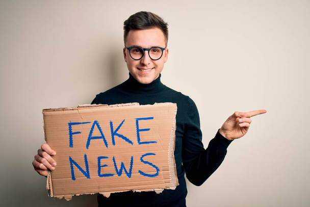 Jovem caucasiano bonito segurando cartão de protesto para desinformação e notícias falsas muito feliz apontando com a mão e o dedo para o lado
 - Foto, Imagem