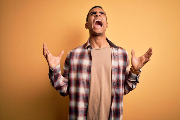 Junger gutaussehender afrikanisch-amerikanischer Mann in lässigem Hemd vor gelbem Hintergrund, verrückt und verrückt schreiend und schreiend mit aggressivem Gesichtsausdruck und erhobenen Armen. Frustrationskonzept. - Foto, Bild