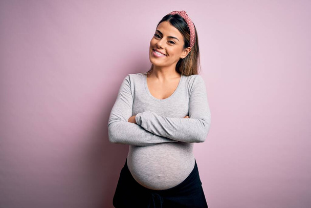 Giovane bella donna bruna incinta in attesa di bambino su sfondo rosa isolato volto felice sorridente con le braccia incrociate guardando la fotocamera. Persona positiva
. - Foto, immagini
