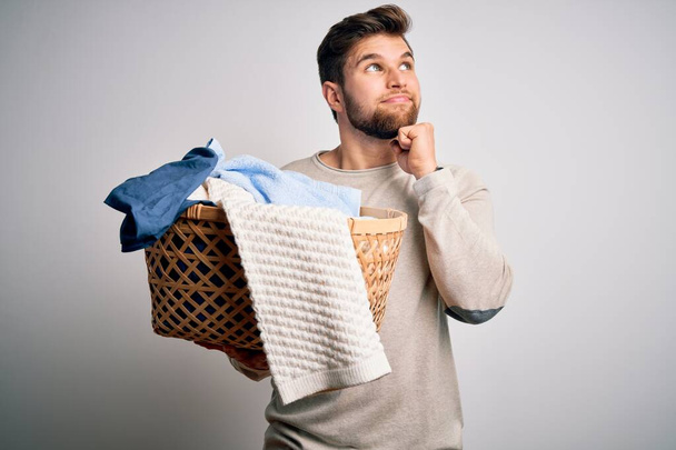 Young blond man with beard and blue eyes doing chores holding wicker basket with clothes serious face thinking about question, very confused idea - Photo, Image