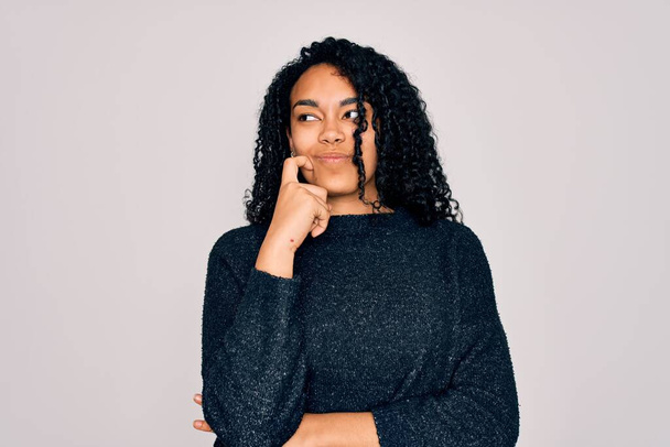 Young african american curly woman wearing casual sweater standing over white background with hand on chin thinking about question, pensive expression. Smiling with thoughtful face. Doubt concept. - Φωτογραφία, εικόνα
