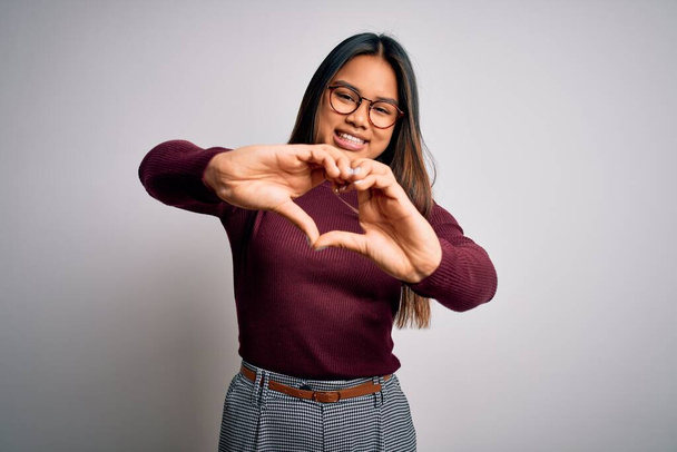 Beautiful asian business woman wearing casual sweater and glasses over white background smiling in love doing heart symbol shape with hands. Romantic concept. - Фото, зображення