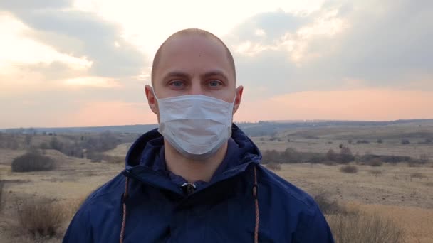 Caucasian young man with medical protective face mask looking to camera standing in the empty field. - Video, Çekim