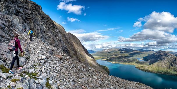 Three young girls with backpacks in bright clothes walking on a rocky slope down to the lake in Norway - Fotoğraf, Görsel