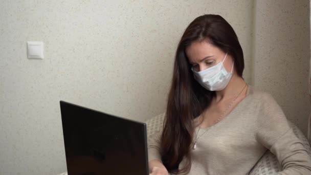 Evde dizüstü bilgisayarla çalışan serbest çalışan genç bir kız. Koruyucu tıbbi yüz maskeli esmer kadın bej bluzlu sandalyede oturup klavyeye yazı yazarak internette sörf yapıyor. Salgın günlerde uzak çalışma. - Video, Çekim