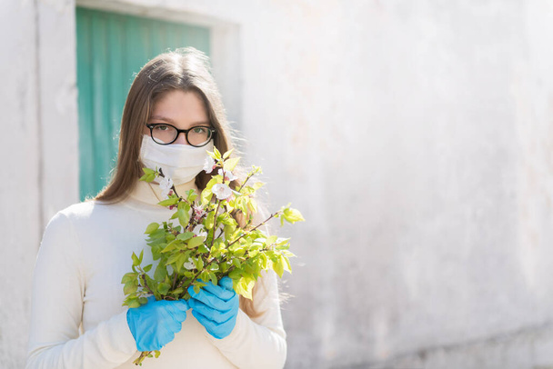 Girl wearing medical mask, is looking with hope and holding a branch of blooming twigs of apricot. Concept of end of quarantine and win over coronavirus and returning to normal life, copy space - Foto, immagini